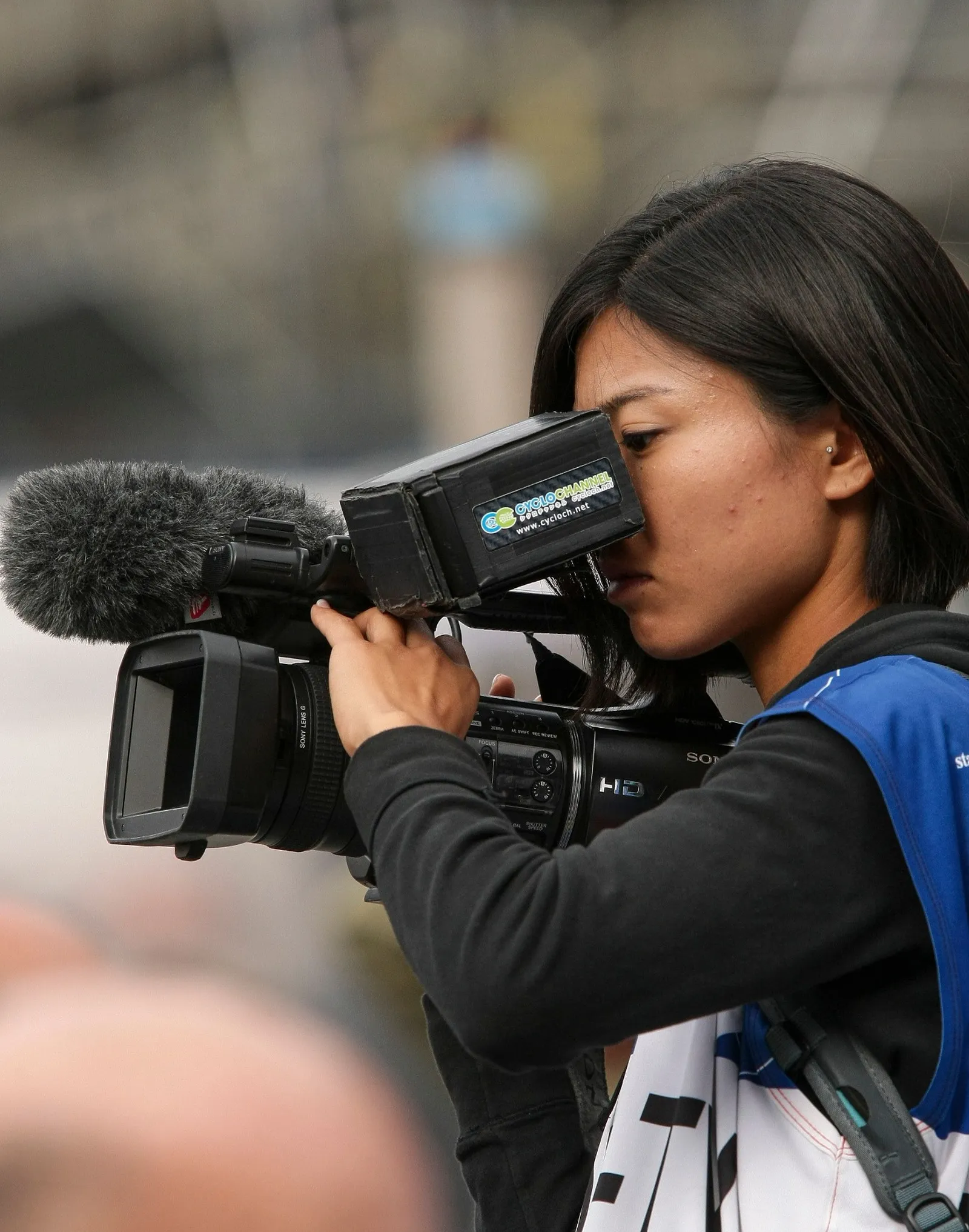 A videographer looks through the viewfinder of a handheld camera outdoors.