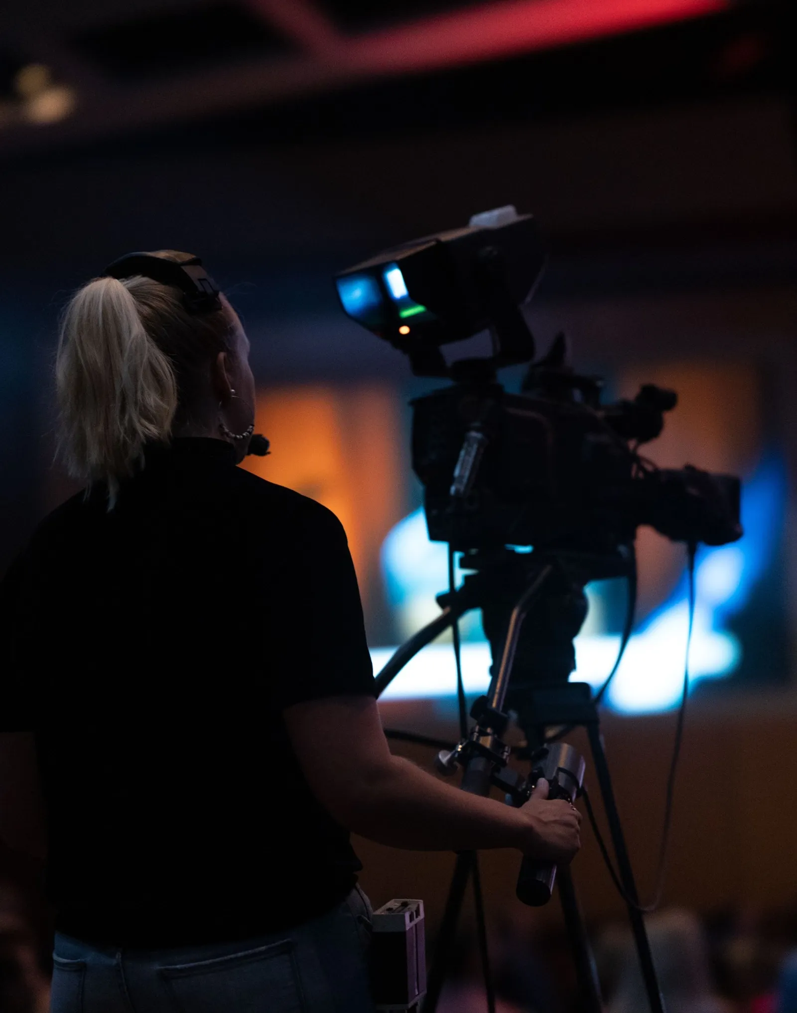 A camera operator wearing a headset films an event from behind a studio camera.