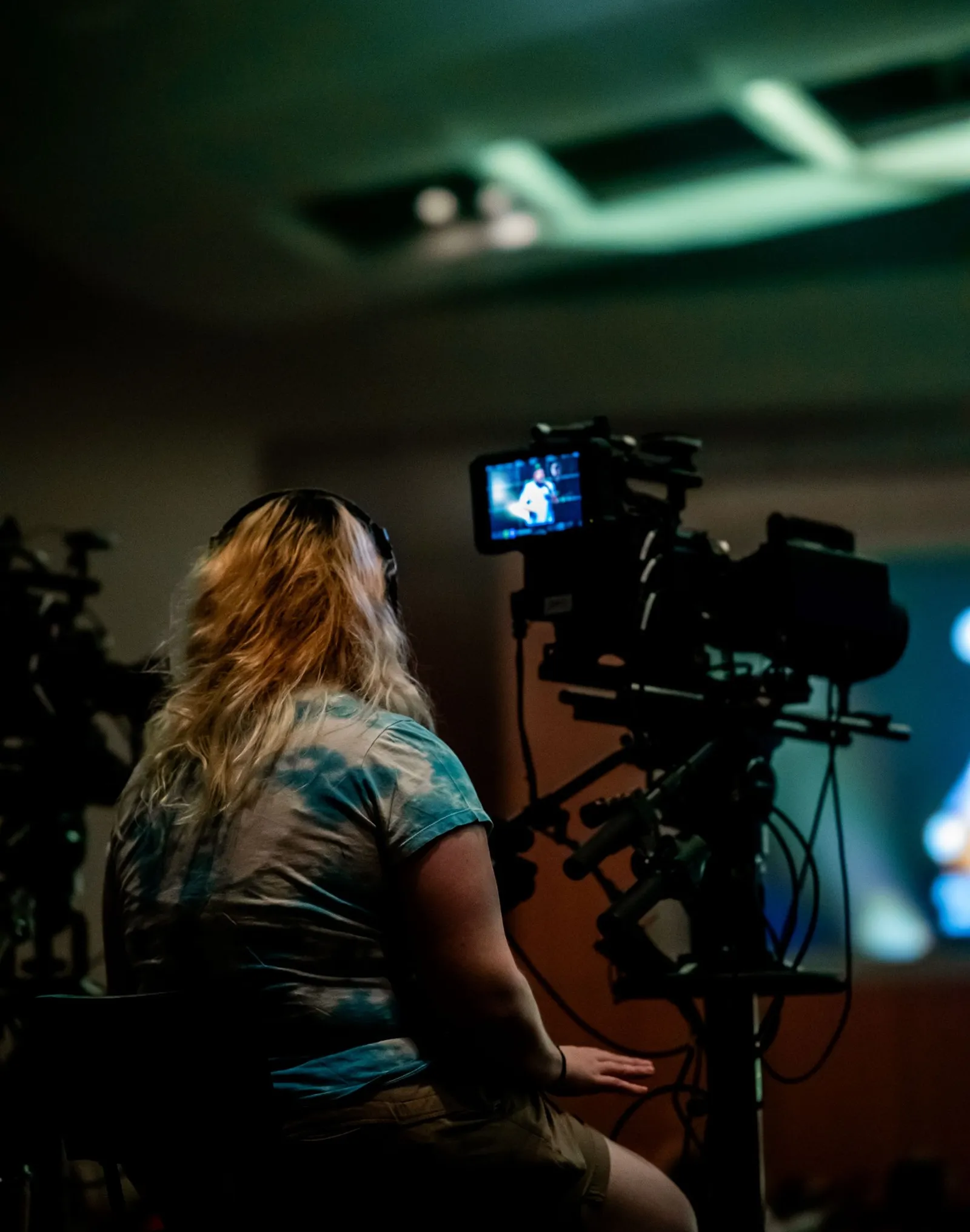 A camera operator seated behind a broadcast camera watches its monitor in a darkened hall.